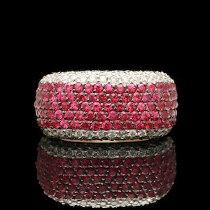 May include: A wide, eye-catching ring with a band of vibrant red gemstones, bordered by rows of sparkling white stones. The ring's design is reflected in the glossy black surface beneath it, creating a symmetrical effect.