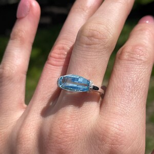 K&ouml;nnte beinhalten: Ein rechteckiger, hellblauer Edelsteinring mit einem silbernen Band. Der Edelstein ist horizontal gesetzt und wird von kleinen, klaren Akzentsteinen flankiert. Der Ring wird an einer Hand mit rosa Nagellack gezeigt.
