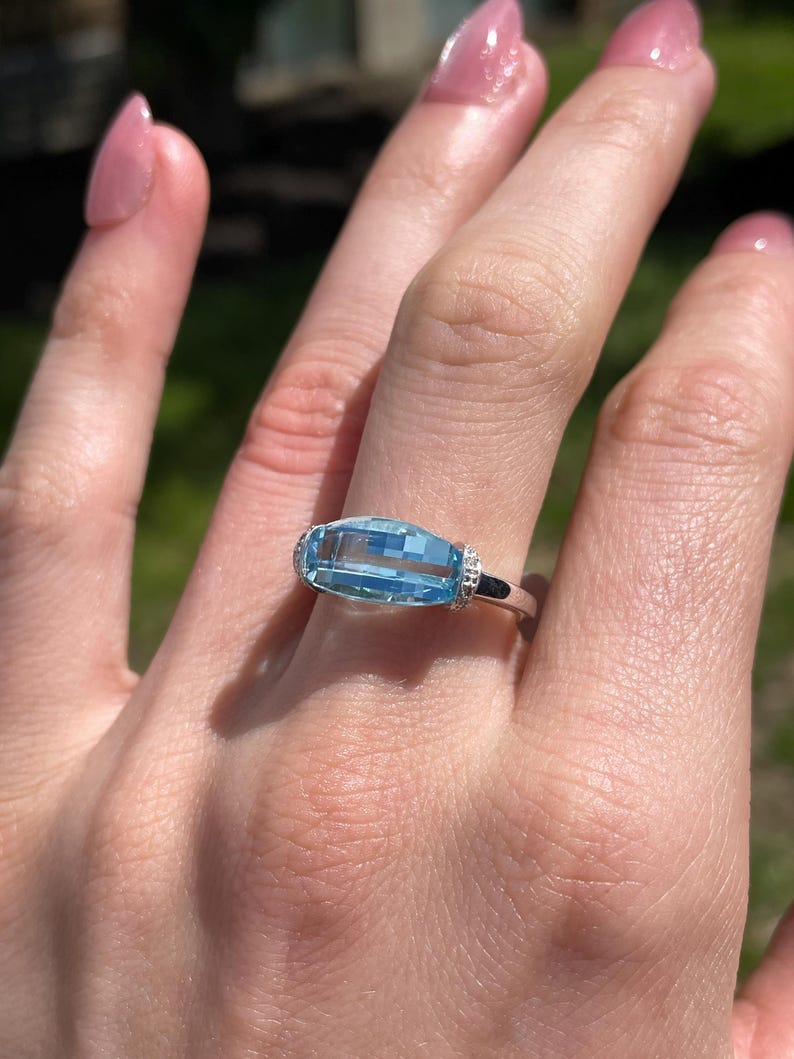 K&ouml;nnte beinhalten: Ein rechteckiger, hellblauer Edelsteinring mit einem silbernen Band. Der Edelstein ist horizontal gesetzt und wird von kleinen, klaren Akzentsteinen flankiert. Der Ring wird an einer Hand mit rosa Nagellack gezeigt.