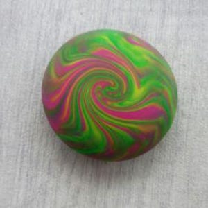 May include: A circular object featuring a vibrant, multi-colored swirl pattern. The design consists of blended streaks of bright lime green, magenta pink, and deep orange, creating a continuous, radiating spiral effect across the entire surface.