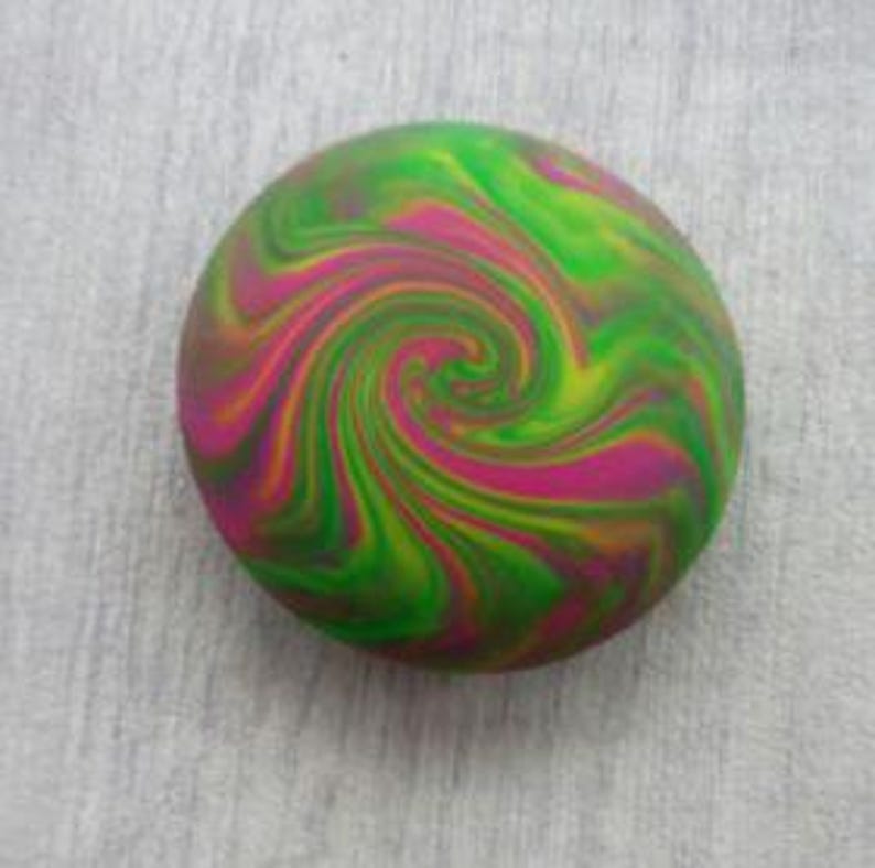 May include: A circular object featuring a vibrant, multi-colored swirl pattern. The design consists of blended streaks of bright lime green, magenta pink, and deep orange, creating a continuous, radiating spiral effect across the entire surface.