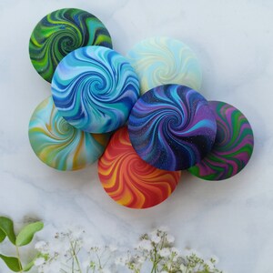 May include: A collection of seven circular objects featuring vibrant, multi-colored swirl patterns. Each piece displays a unique color combination, including shades of green, blue, purple, orange, and yellow, with some designs incorporating fine glitter accents.