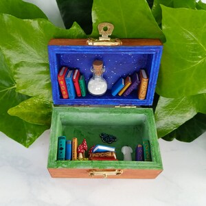 May include: A wooden box with a hinged lid, painted in shades of brown, blue, and green. The box is open, revealing a miniature scene with books, a bottle, mushrooms, and a butterfly. The box has a metal latch and a keyhole.
