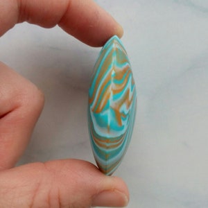 May include: A smooth, almond-shaped object featuring a marbled pattern of turquoise, white, and tan swirls. The piece has a pointed top and bottom with a rounded, convex profile, displaying a continuous, fluid design across its entire surface.