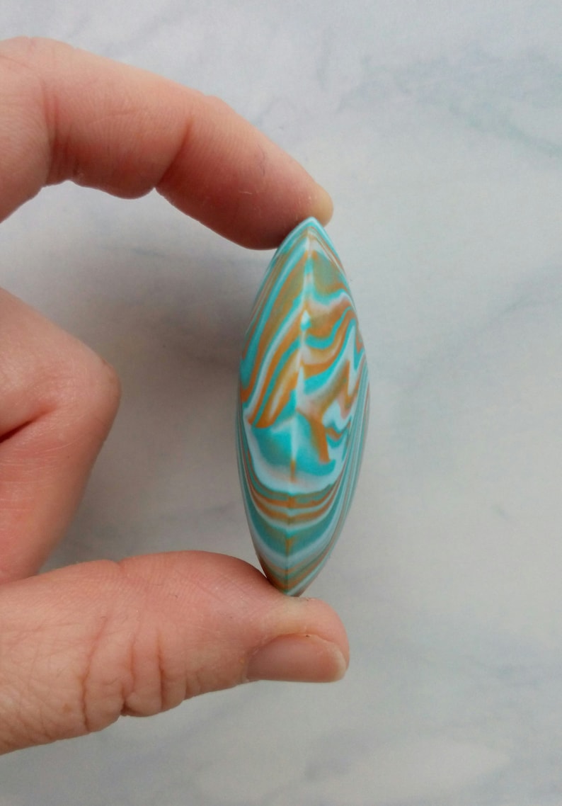 May include: A smooth, almond-shaped object featuring a marbled pattern of turquoise, white, and tan swirls. The piece has a pointed top and bottom with a rounded, convex profile, displaying a continuous, fluid design across its entire surface.