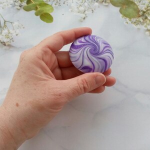 May include: A round, smooth object featuring a swirling pattern of deep purple, light lavender, and white colors. The surface has a subtle glittery finish that catches the light, creating a marbled effect across the circular, flat-topped shape.
