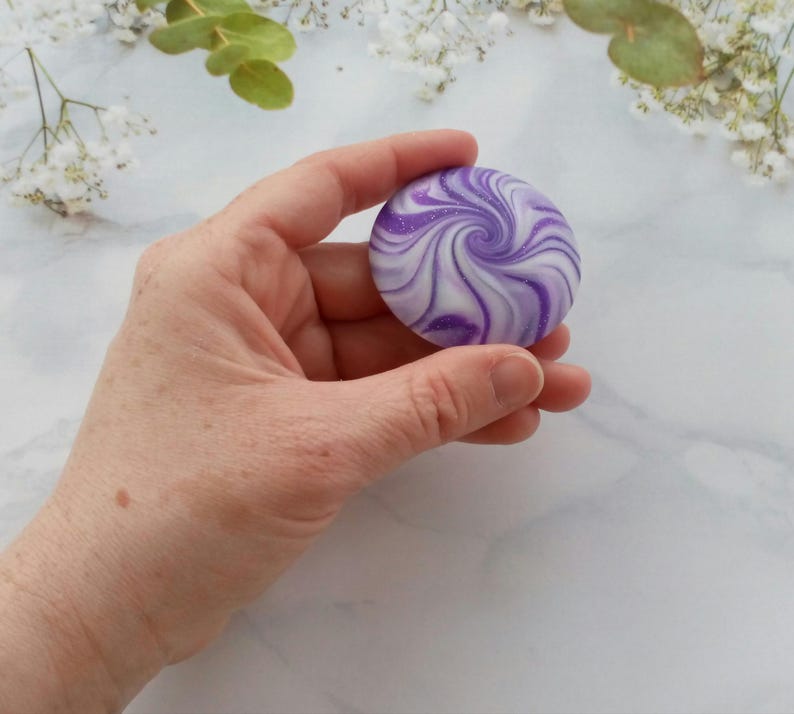 May include: A round, smooth object featuring a swirling pattern of deep purple, light lavender, and white colors. The surface has a subtle glittery finish that catches the light, creating a marbled effect across the circular, flat-topped shape.
