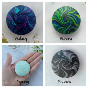 May include: Four circular objects with swirling patterns. The Galaxy design features blue, purple, and black with glitter. The Aurora design has green and blue swirls. The Spring design is light blue and yellow. The Shadow design is black and grey.
