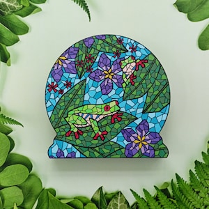 May include: A green tree frog with red eyes sits on a mosaic tile with blue, green, and purple flowers. The mosaic tile is surrounded by green leaves.