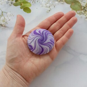 May include: A circular, flat object featuring a swirling pattern of deep purple and white colors. The surface has a smooth, polished appearance with subtle glittery accents integrated into the purple swirls, creating a marbled effect.