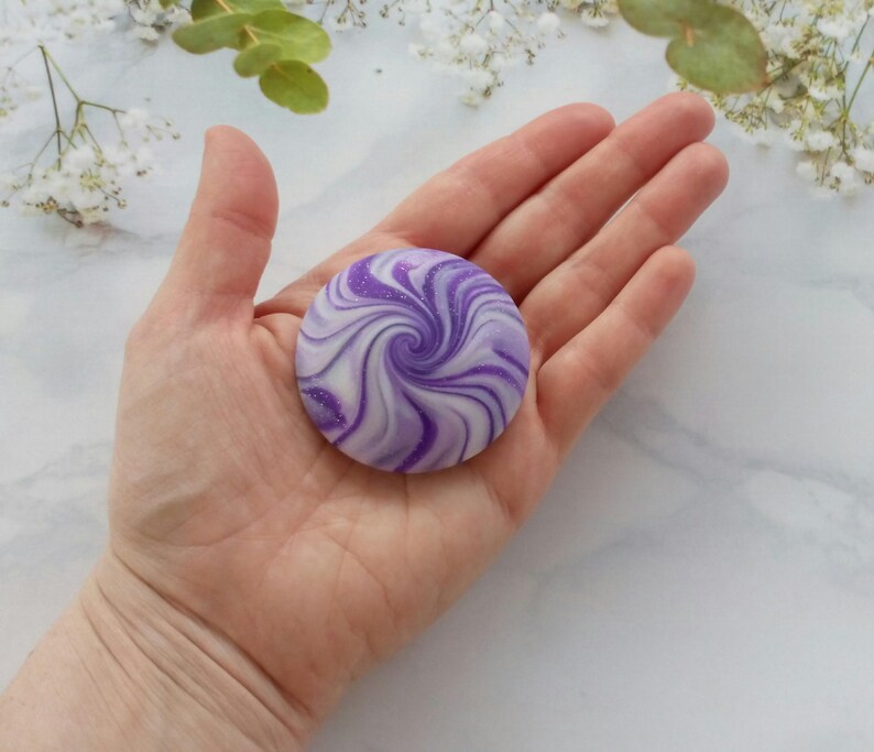 May include: A circular, flat object featuring a swirling pattern of deep purple and white colors. The surface has a smooth, polished appearance with subtle glittery accents integrated into the purple swirls, creating a marbled effect.