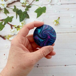 May include: A circular object featuring a swirling galaxy pattern in shades of deep blue, purple, and teal. The surface is decorated with fine white speckles resembling distant stars, creating a cosmic effect across the round, smooth disc.