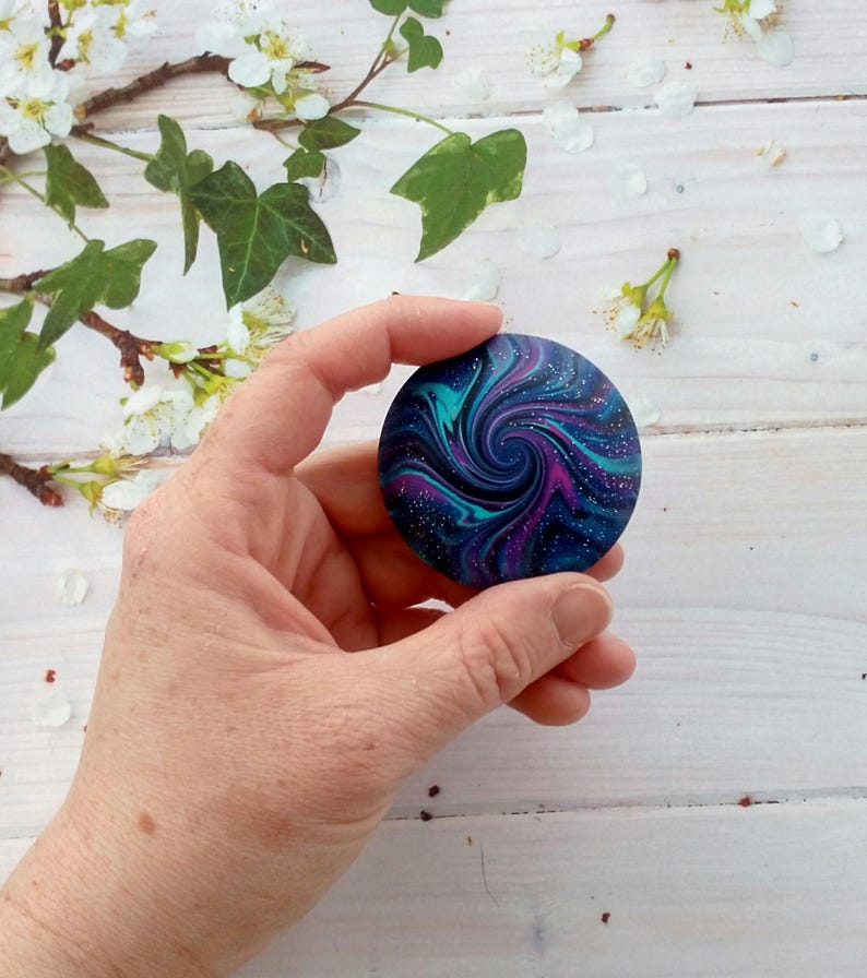 May include: A circular object featuring a swirling galaxy pattern in shades of deep blue, purple, and teal. The surface is decorated with fine white speckles resembling distant stars, creating a cosmic effect across the round, smooth disc.