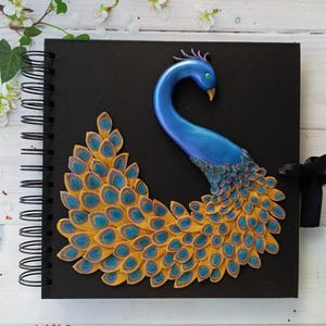 May include: Black spiral-bound journal with a 3D peacock design. The peacock features a blue body and a fan of yellow, blue, and purple feathers. A black ribbon is tied on the right side. The journal is a decorative item.