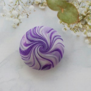 May include: A circular object featuring a swirling pattern of deep purple, light lavender, and white colors. The surface has a smooth finish with fine, scattered glitter particles embedded throughout the design, creating a spiral effect.