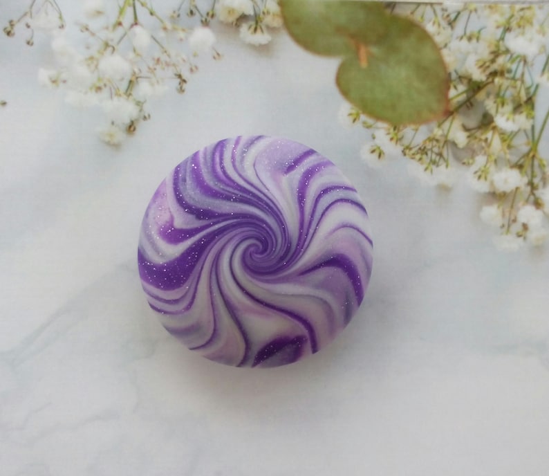 May include: A circular object featuring a swirling pattern of deep purple, light lavender, and white colors. The surface has a smooth finish with fine, scattered glitter particles embedded throughout the design, creating a spiral effect.