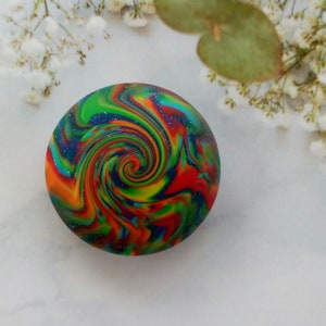 May include: A circular object featuring a vibrant, multi-colored swirl pattern with streaks of red, orange, yellow, green, and blue. The surface is embedded with fine, reflective glitter particles that create a sparkling effect throughout the design.