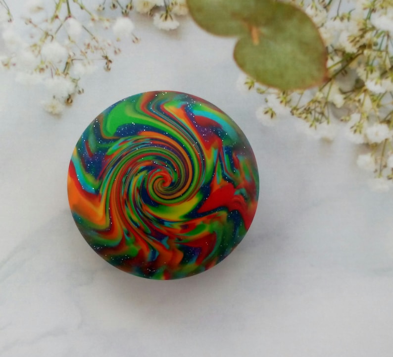 May include: A circular object featuring a vibrant, multi-colored swirl pattern with streaks of red, orange, yellow, green, and blue. The surface is embedded with fine, reflective glitter particles that create a sparkling effect throughout the design.