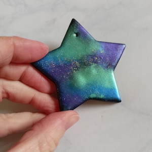 May include: A star-shaped ornament with a hole at the top. The ornament features a blend of blue, green, and purple hues with gold flecks. The edges are black, and the star is held by a hand.