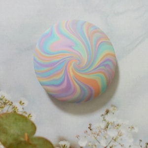 May include: A circular, flat object featuring a swirling, marbled pattern of pastel colors including light blue, soft pink, lavender, and peach. The design radiates from the center in a continuous, smooth, and blended spiral motion.