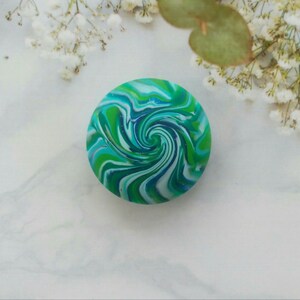 May include: A circular object featuring a vibrant marbled pattern of swirling blue, teal, lime green, and white colors. The surface has a smooth, glossy finish with subtle glitter accents integrated into the colorful, fluid, and hypnotic spiral design.