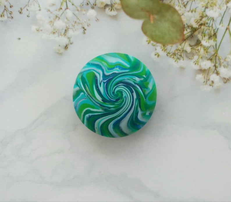 May include: A circular object featuring a vibrant marbled pattern of swirling blue, teal, lime green, and white colors. The surface has a smooth, glossy finish with subtle glitter accents integrated into the colorful, fluid, and hypnotic spiral design.