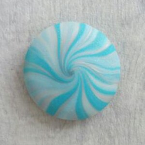 May include: A circular button featuring a swirling pattern of bright turquoise and white colors. The design radiates from the center outward in a spiral motion, creating a smooth, rounded surface with a consistent color distribution throughout.