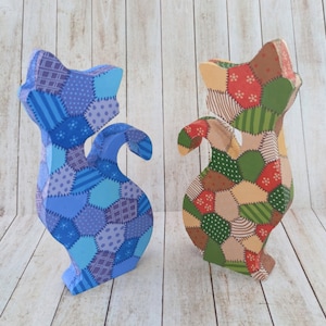 May include: Two wooden cat figurines, one blue and one green, with a patchwork design. The cats are standing on a light wood background.