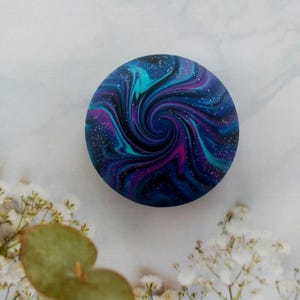 May include: A circular object featuring a swirling galaxy pattern in shades of deep blue, vibrant purple, and bright teal. The surface is speckled with tiny white dots that resemble stars, creating a cosmic effect across the entire round face.