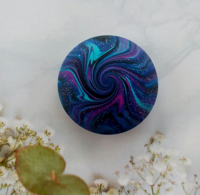 May include: A circular object featuring a swirling galaxy pattern in shades of deep blue, vibrant purple, and bright teal. The surface is speckled with tiny white dots that resemble stars, creating a cosmic effect across the entire round face.