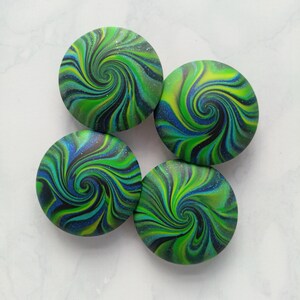 May include: Four circular buttons featuring a swirling marbled pattern in shades of lime green, deep blue, and black. Each button has a smooth, glossy finish with fine glitter particles embedded throughout the surface, creating a vibrant, multi-colored effect.