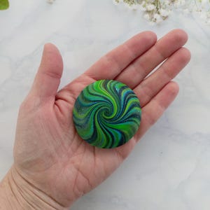 May include: A circular, flat object featuring a vibrant swirl pattern in shades of lime green, forest green, teal, and deep navy blue. The surface has a subtle, shimmering glitter finish that catches the light, creating a dynamic, spiraling effect.