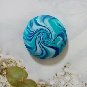 May include: A circular cabochon featuring a marbled swirl pattern in shades of turquoise, royal blue, and white. The surface has a smooth, glossy finish with subtle embedded glitter particles that catch the light, creating a dynamic spiral effect.