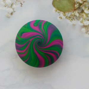 May include: A circular object featuring a vibrant, swirling pattern of magenta, bright green, and deep blue colors. The design radiates from the center in a continuous, fluid spiral motion, creating a bold and colorful aesthetic on the round surface.
