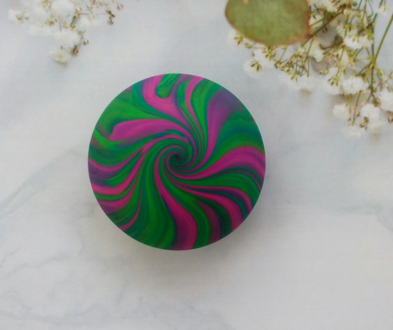 May include: A circular object featuring a vibrant, swirling pattern of magenta, bright green, and deep blue colors. The design radiates from the center in a continuous, fluid spiral motion, creating a bold and colorful aesthetic on the round surface.