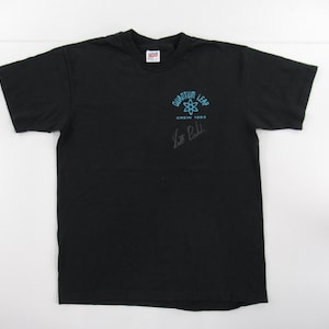Puede incluir: Camiseta negra con un logotipo de Quantum Leap azul y blanco y el texto "Crew 1993". La camiseta est&aacute; firmada por "Scott Bakula".