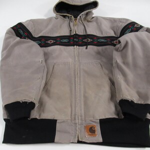 May include: A gray hooded jacket with a black and brown patterned band across the chest. The jacket has a zipper closure and a Carhartt logo patch on the bottom right side.