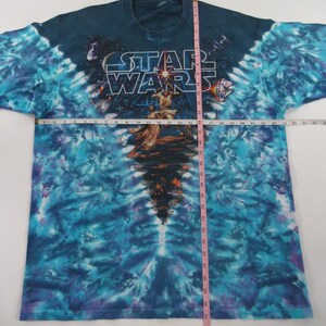 Puede incluir: Una camiseta tie-dye azul, morado y blanco con un gr&aacute;fico de Star Wars. El gr&aacute;fico presenta a Luke Skywalker, Darth Vader y el Halc&oacute;n Milenario. El texto "Star Wars" est&aacute; impreso en blanco.