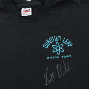 Puede incluir: Camiseta negra con un logotipo de Quantum Leap azul y blanco, que presenta un s&iacute;mbolo de &aacute;tomo y el texto "Crew 1993". La camiseta est&aacute; firmada por Scott Bakula.