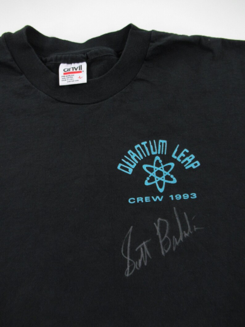 Puede incluir: Camiseta negra con un logotipo de Quantum Leap azul y blanco, que presenta un s&iacute;mbolo de &aacute;tomo y el texto "Crew 1993". La camiseta est&aacute; firmada por Scott Bakula.