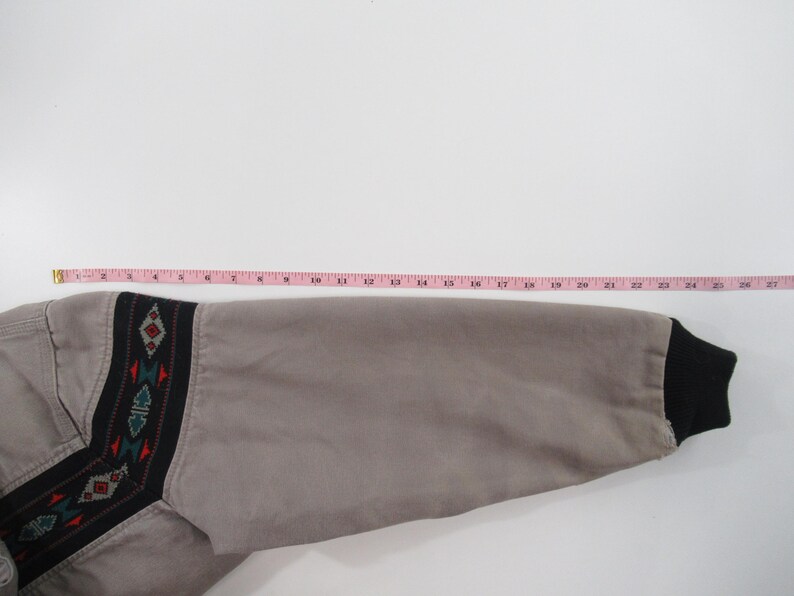 May include: A gray jacket with a black and brown geometric pattern on the sleeve. The sleeve is rolled up and the cuff is black.