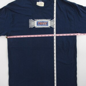Op de afbeelding: Een marineblauw T-shirt met een witte en blauwe afbeelding van een computerchip met het woord "NIKE" in rode en witte letters. Het shirt wordt gemeten met een meetlint, waarbij de lengte 68,58 cm is.