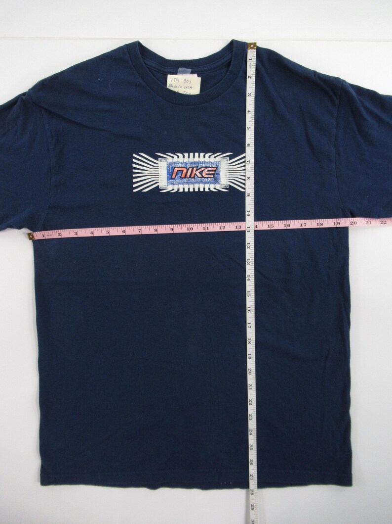 Op de afbeelding: Een marineblauw T-shirt met een witte en blauwe afbeelding van een computerchip met het woord "NIKE" in rode en witte letters. Het shirt wordt gemeten met een meetlint, waarbij de lengte 68,58 cm is.