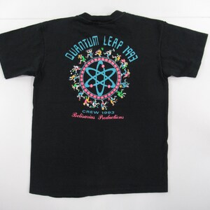 Puede incluir: Camiseta negra con un gr&aacute;fico colorido de un c&iacute;rculo con un s&iacute;mbolo de &aacute;tomo en su interior y el texto "Quantum Leap 1993" encima. El texto "Crew 1993 Belisarius Productions" est&aacute; debajo del c&iacute;rculo.