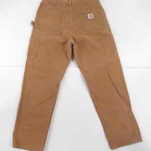 Puede incluir: Un par de pantalones de trabajo de mezclilla marr&oacute;n con un parche de logotipo de Carhartt en el bolsillo trasero derecho.