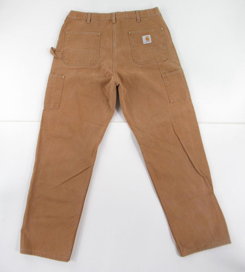 Puede incluir: Un par de pantalones de trabajo de mezclilla marr&oacute;n con un parche de logotipo de Carhartt en el bolsillo trasero derecho.