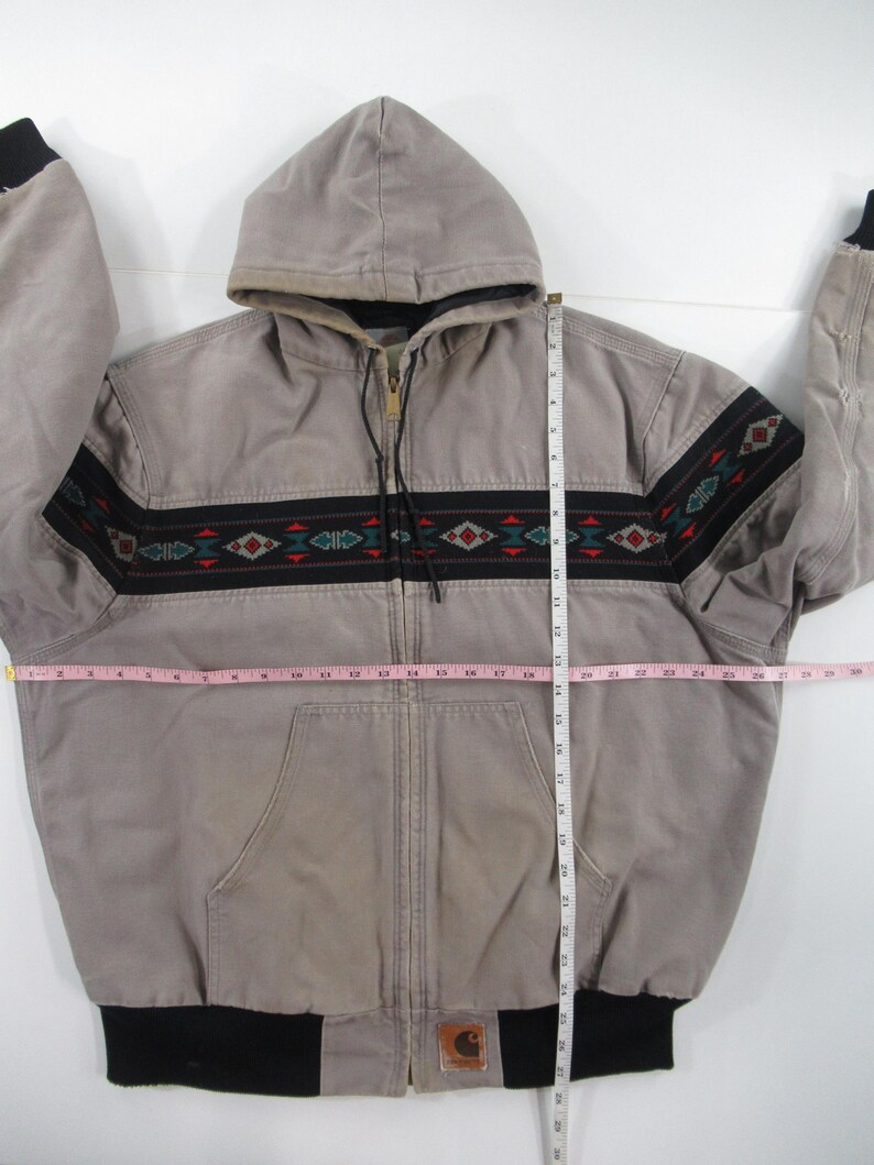 May include: A gray hooded Carhartt jacket with a black and red patterned band around the waist. The jacket has a zipper closure and a black Carhartt logo patch on the bottom right side.