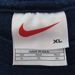 Op de afbeelding: Een wit label met een rood Nike swoosh-logo en de tekst "XL" in zwart. Het label heeft ook de tekst "Made in U.S.A." en maat informatie in het Engels, Frans en Spaans.