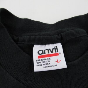 Puede incluir: Una camiseta negra con una etiqueta blanca que dice "Anvil" y "Pre-Shrunk 100% Cotton Made in U.S.A. Over for Care"