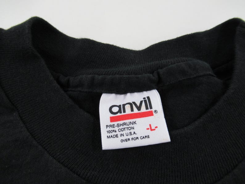 Puede incluir: Una camiseta negra con una etiqueta blanca que dice "Anvil" y "Pre-Shrunk 100% Cotton Made in U.S.A. Over for Care"